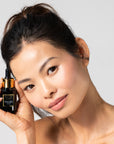 Woman holding a Black serum bottle with dropper labeled 'Bella Vida Santa Barbara Sunlight Serum with Acetyl Hexapeptide-8 and Bakuchiol