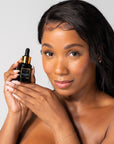 Woman holding a Black serum bottle with dropper labeled 'Bella Vida Santa Barbara Sunlight Serum with Acetyl Hexapeptide-8 and Bakuchiol