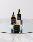 Three black Bella Vida Santa Barbara luxury skincare bottles with gold caps on a round glass table. Sunlight Serum, Cocomero Watermelon Glow Serum and Brighten Vitamin C Cream
